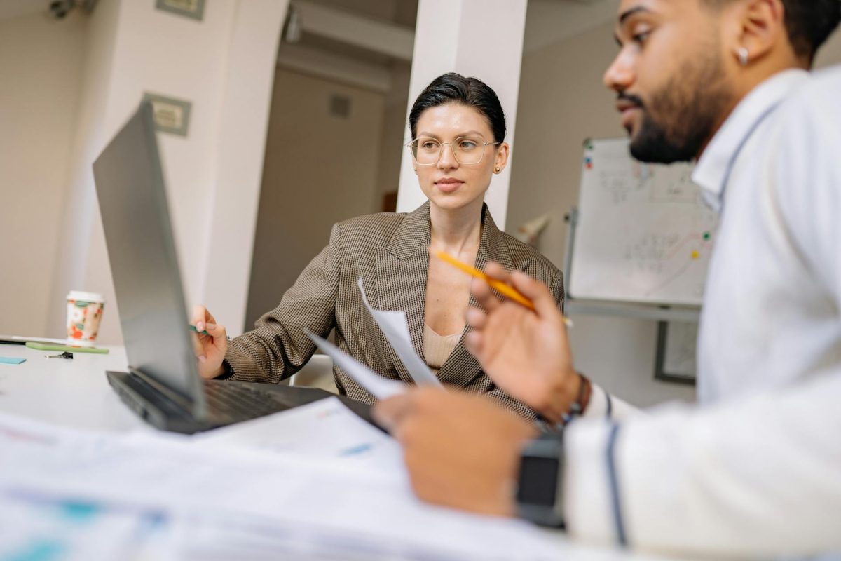 A man and a woman holding pencils, looking at a laptop, discussing an online business exit strategy.